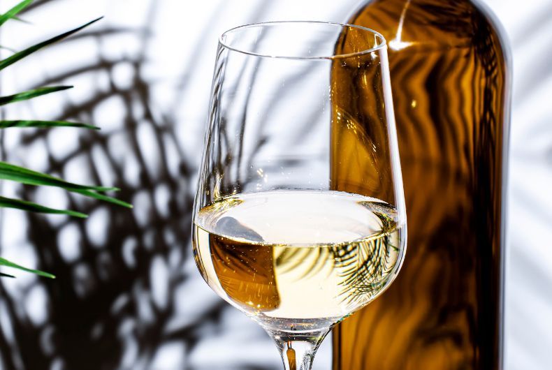 Vino varietale Valtidone: primo piano di un bicchiere di vino bianco con bottiglia ambrata sullo sfondo e ombre di foglie.