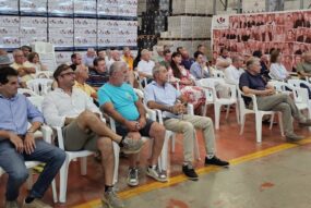 Gruppo di persone sedute su sedie bianche in un magazzino industriale con pile di scatole di vino e un pannello fotografico Cantina Valtidone.