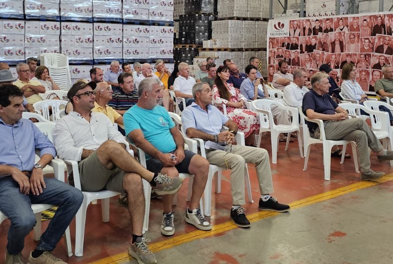 Gruppo di persone sedute su sedie bianche in un magazzino industriale con pile di scatole di vino e un pannello fotografico Cantina Valtidone.